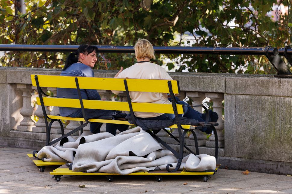 A photo of two people sitting on a bright yellow bench on a terrace with a stone railing. Rolled-up fabric lies under the bench, and trees provide shade in the background.