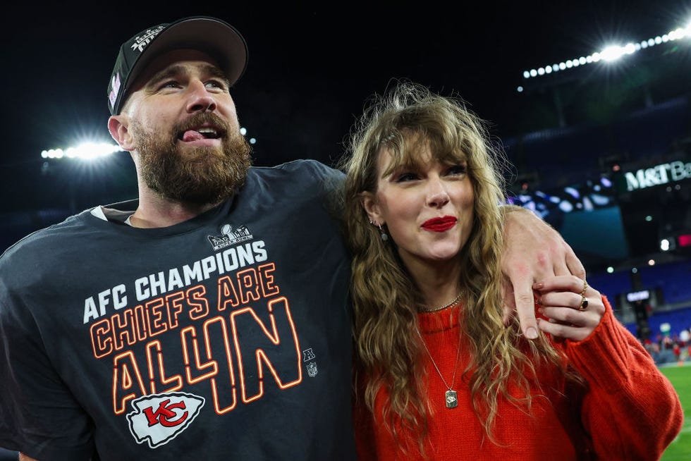 travis kelce and taylor swift stand on a football field at night, he has one arm around her shoulders and they hold hands