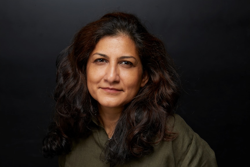 Nusra Latif Qureshi, with long brown hair and dark green top, seen from waist up, smiling with mouth closed.