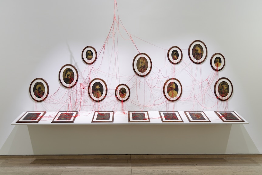 In a white room, a display cabinet shows several framed paintings just visible, and above it connected by red string are more.