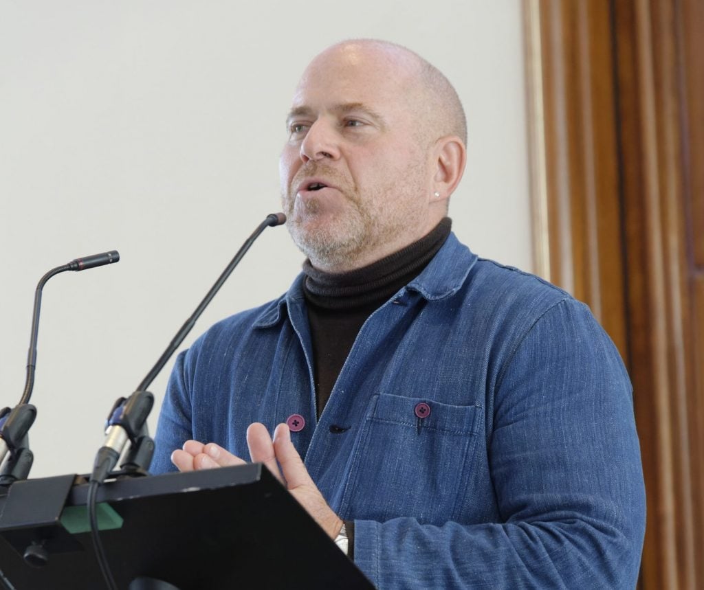 A caucasian man with skin head, dressed in a blue shirt over a black turtle neck, speaking on a podium with microphones,