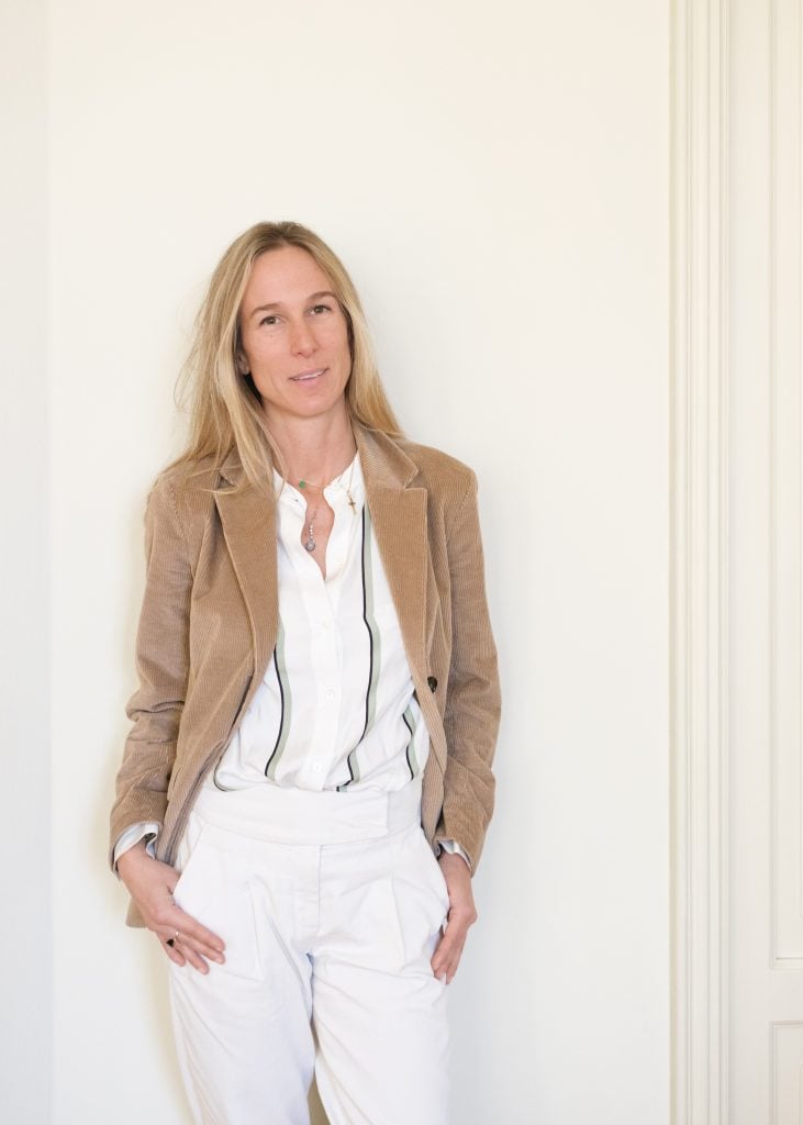 A caucasian woman with long blonde hair, earth tone blazer, white shirt and white trousers.