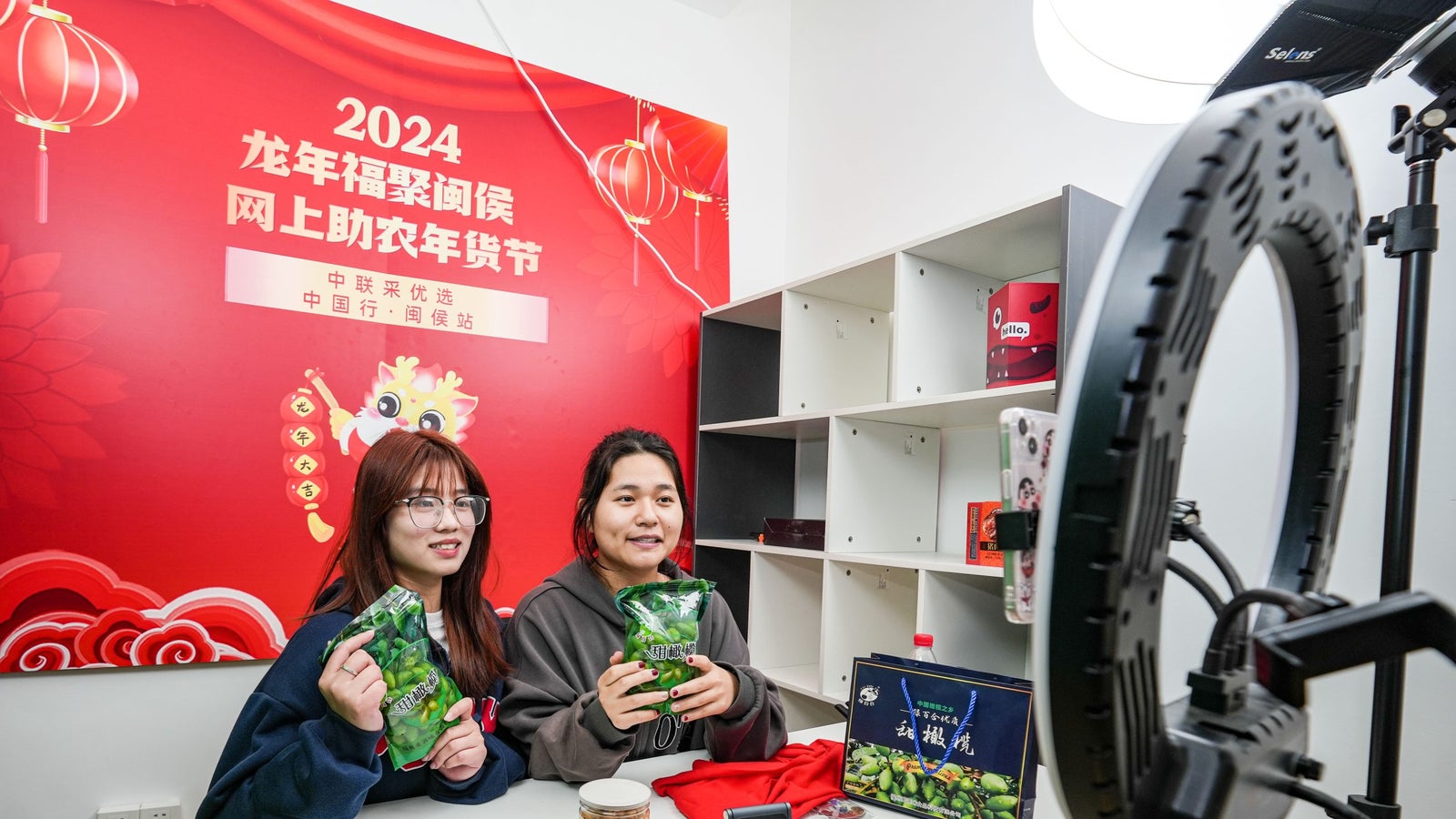 Two women are sitting at a table displaying green snacks, smiling at a camera. They are in front of a colorful backdrop promoting a 2024 event. The table features jars of snacks, a red cloth, and a decorative box. A ring light illuminates the scene.