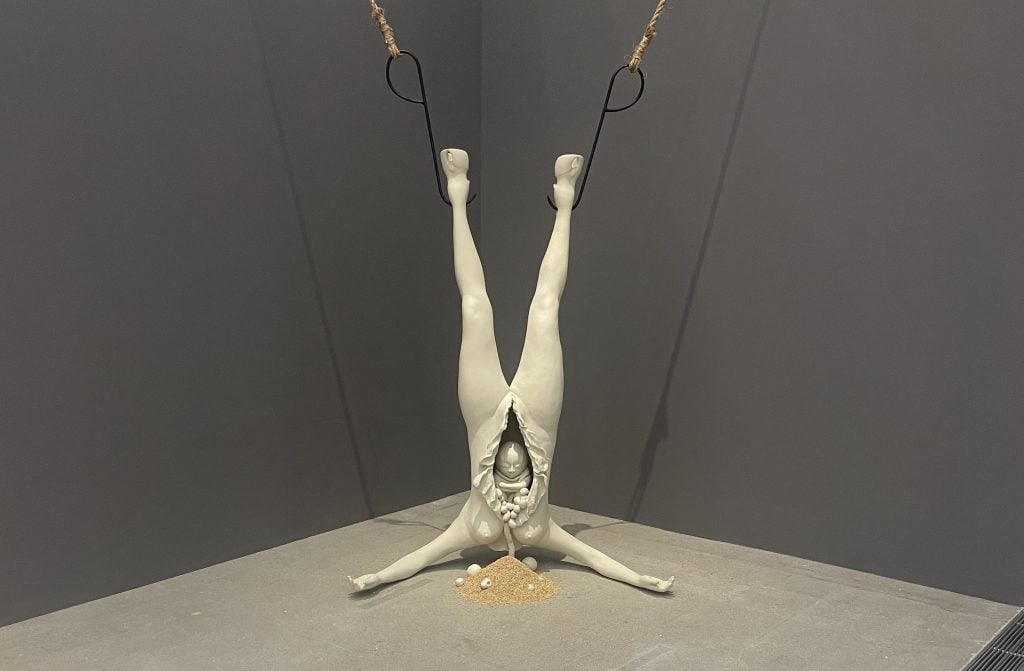 A white sculpture resembling the body of a woman but with horse feet, hung upside down. Her belly is open, revealing a baby sitting inside. 