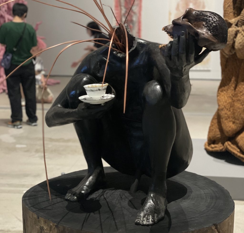 A black sculpture of a headless figure, crouching on a wood stand, holding a white tea cup. Bronze wires grew from the wire's neck. 