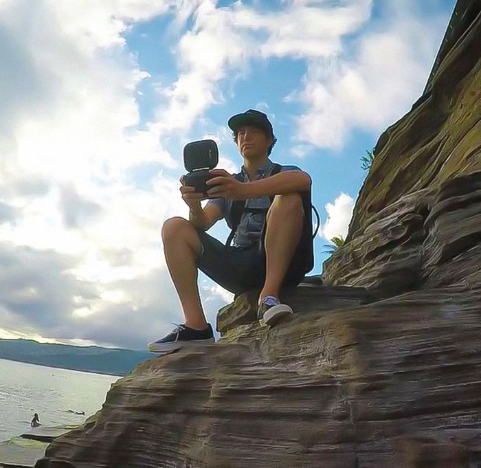 Man sitting on a rocky cliff, holding a camera against a scenic backdrop, reminiscent of MySpace founder.