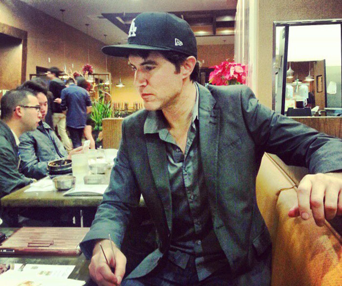 Man in a suit and cap sits at a restaurant booth, writing on paper, representing MySpace founder in casual setting.