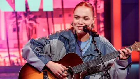 Getty Images UPSAHL performing July 31, 2024 in Miami Beach, Florida. Shehas pulled back reddish hair and is wearing a grey and blue jacket, possibly leather. She is seated and playing a guitar, looking slightly over to her right, with a microphone on a stand in front of her