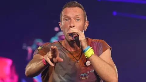 PA Media  Coldplay's Chris Martin performing on stage at Wembley Stadium, north London, in 2022. He has short brown hair and stubble and is holding the microphone up to his mouth with his left hand. His right hand is outstretched. He is wearing a reddish t-shirt