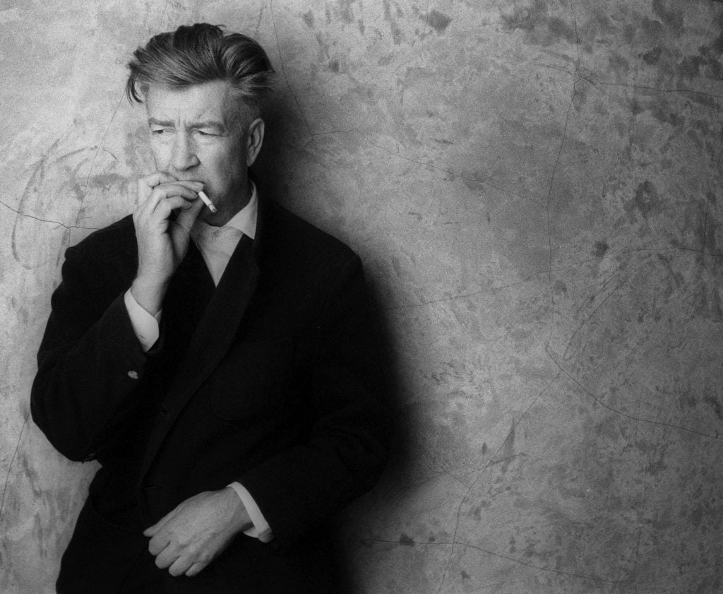 Portrait of David Lynch, holding a cigarette and posing against a stained wall.