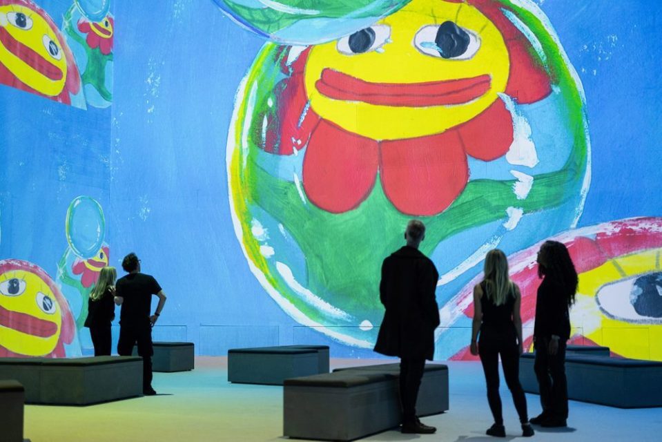 People viewing a large-scale projection of colorful, cartoonish flower-like imagery.