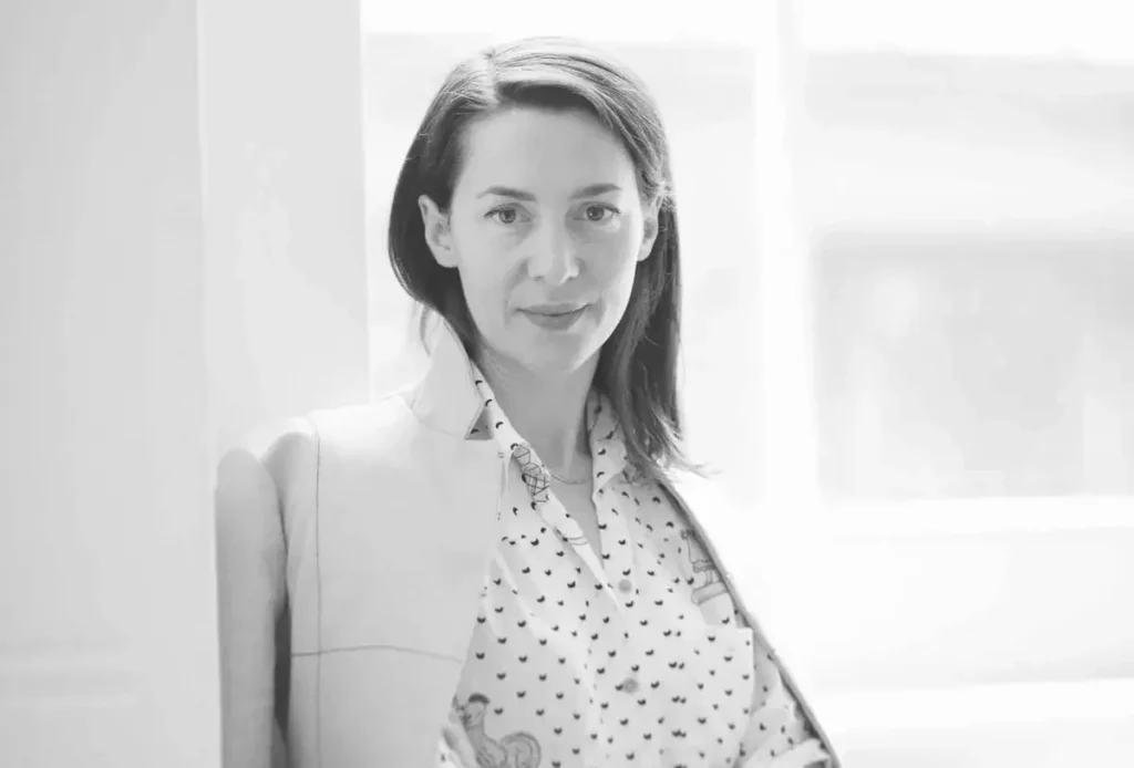 a black and white photo of a woman in a jacket and dotted shirt leaning up against a wall