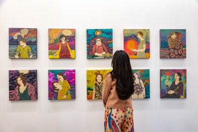 A woman looks at a 5x2 grid of figurative paintings of women in surrealistic setting. 