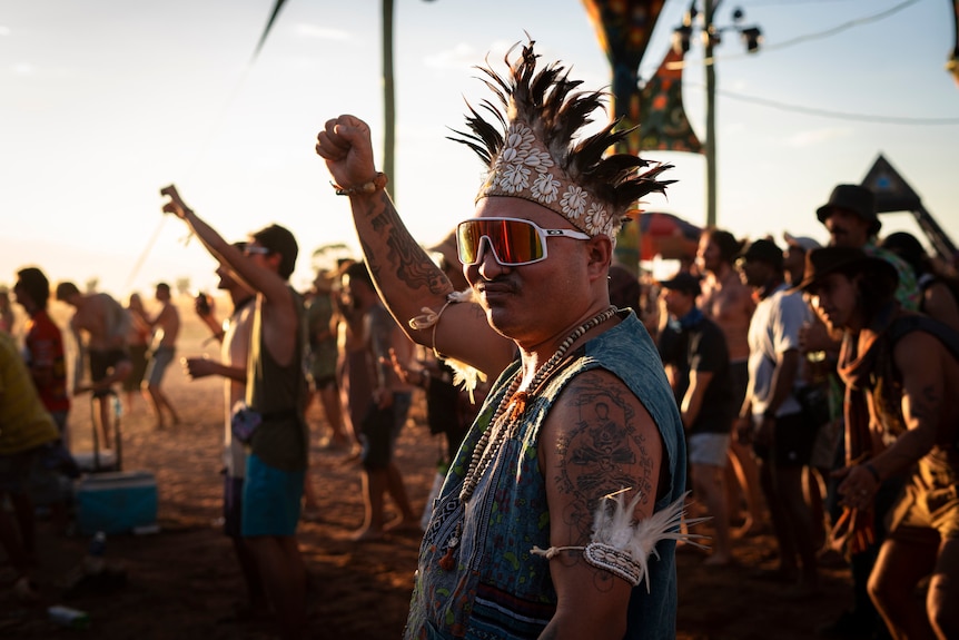 A man in a headdress and goggles raises his fist as he dances at an outdoor music festival.