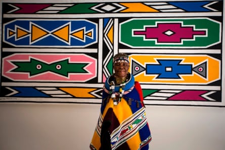 Esther Mahlangu in front of a large Ndebele-style artwork in vibrant geometric patterns and bold primary colours of red, green, yellow and blue, as well as white and pink.