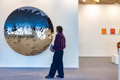 A man looks at a wall-hung sculpture of reflective metal in the shape of a circle. 