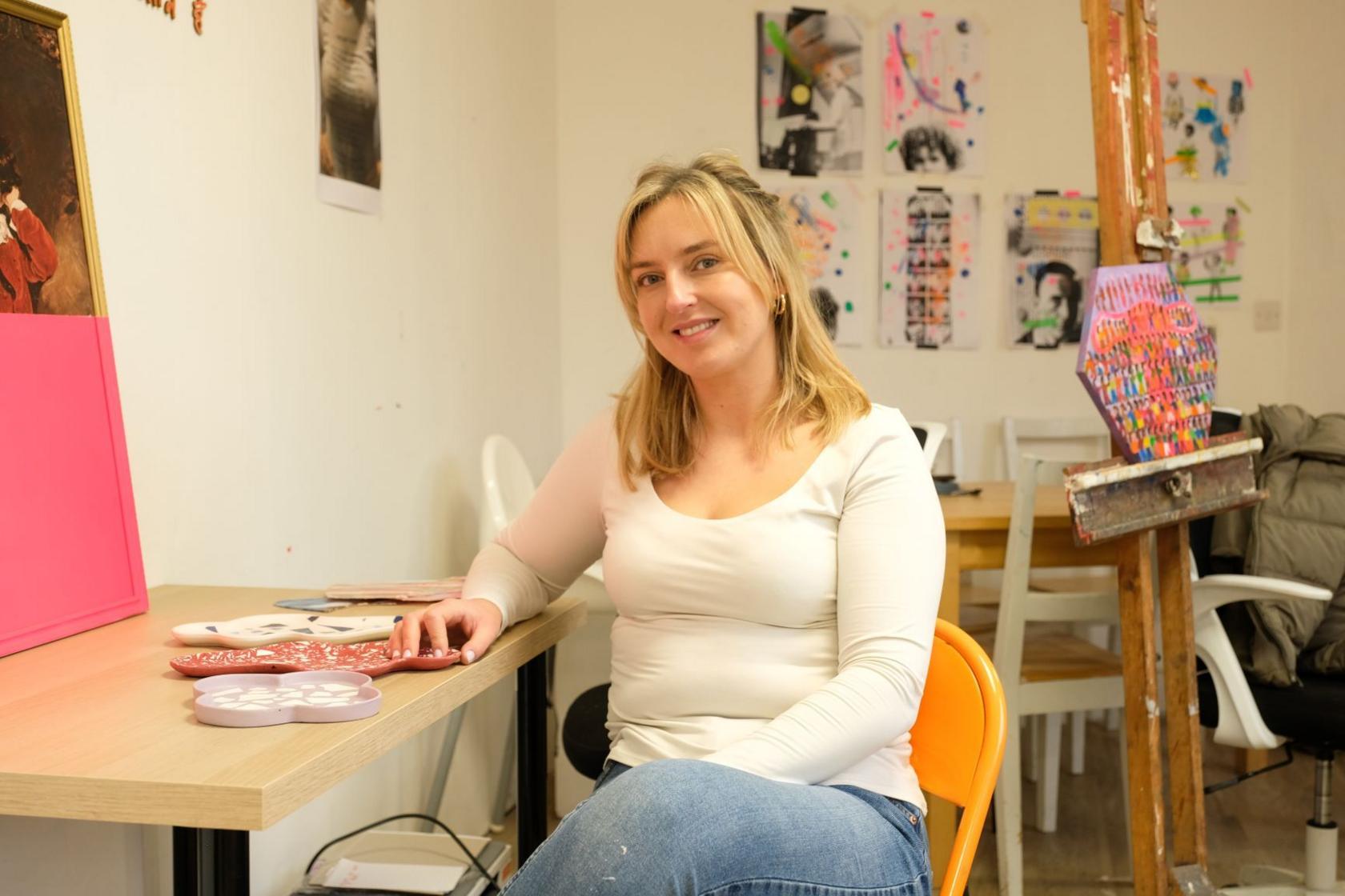 A woman sits at a table in an art studio. She has blonde hair, and she is wearing blue jeans and a white top. There are multi-coloured plaster casts, shaped like dishes, on the table. An easel with a multi-coloured painting can be seen in the background.