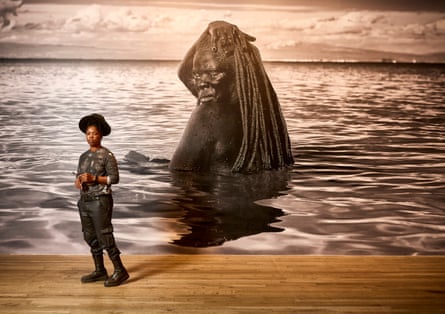 Zanele Muholi standing in front of a large artwork of a black person with dreadlocks bathing in the sea