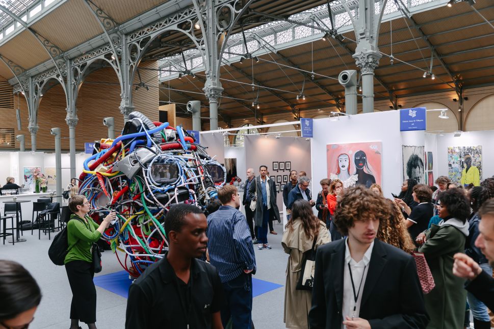 An art fair scene at AKAA (Also Known as Africa) captures a large crowd inside a historic building with ornate ironwork, where people are viewing contemporary African art, including a striking sculpture made of multicolored wires and a painting of two women with dark and light skin.