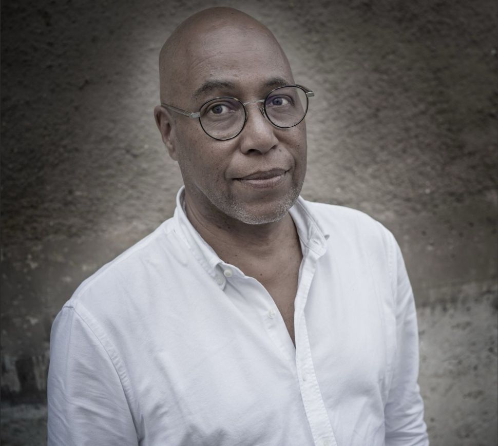 A portrait photograph of Sitor Senghor shows him wearing a white button-down shirt and round glasses, standing against a textured grayish wall, looking directly at the camera with a calm and confident expression; the image is credited to photographer Antoine Tempé.