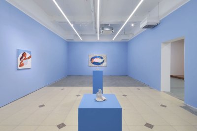 View of an art gallery with blue painted walls, two paintings on the walls, and two sculptures on individual plinth. 