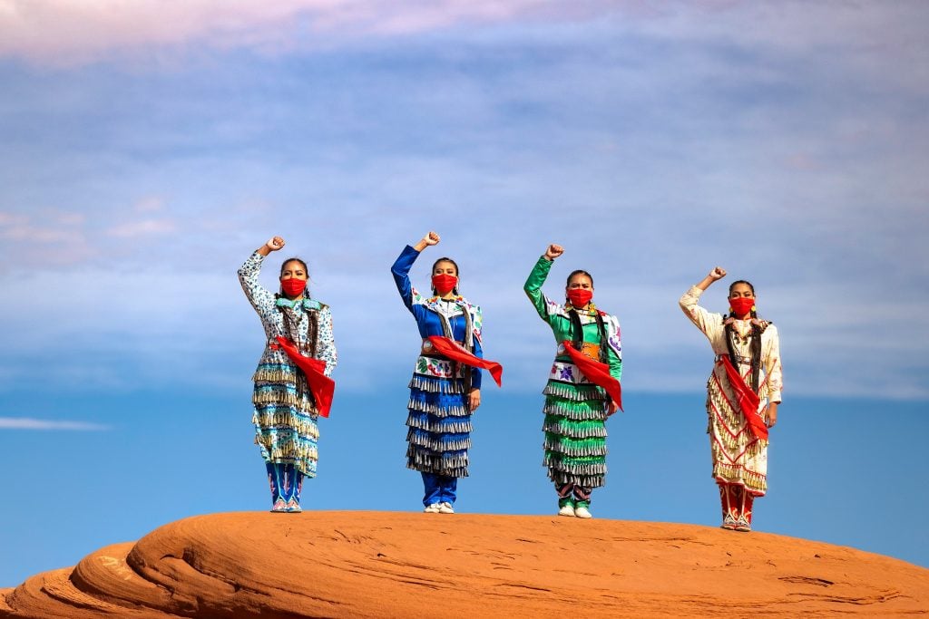 A photograph titled Salaridarity by Eugene Tapahe, exhibited at Monroe Gallery. Four Indigenous women stand atop a red sandstone formation, wearing traditional jingle dresses in vibrant colors—white, blue, green, and patterned designs. Each woman raises her fist in a gesture of strength and solidarity while wearing red face masks and sashes. The background features a vast sky with soft hues of blue and pink, emphasizing the powerful stance of the women. The image is a tribute to Indigenous culture, resilience, and unity.