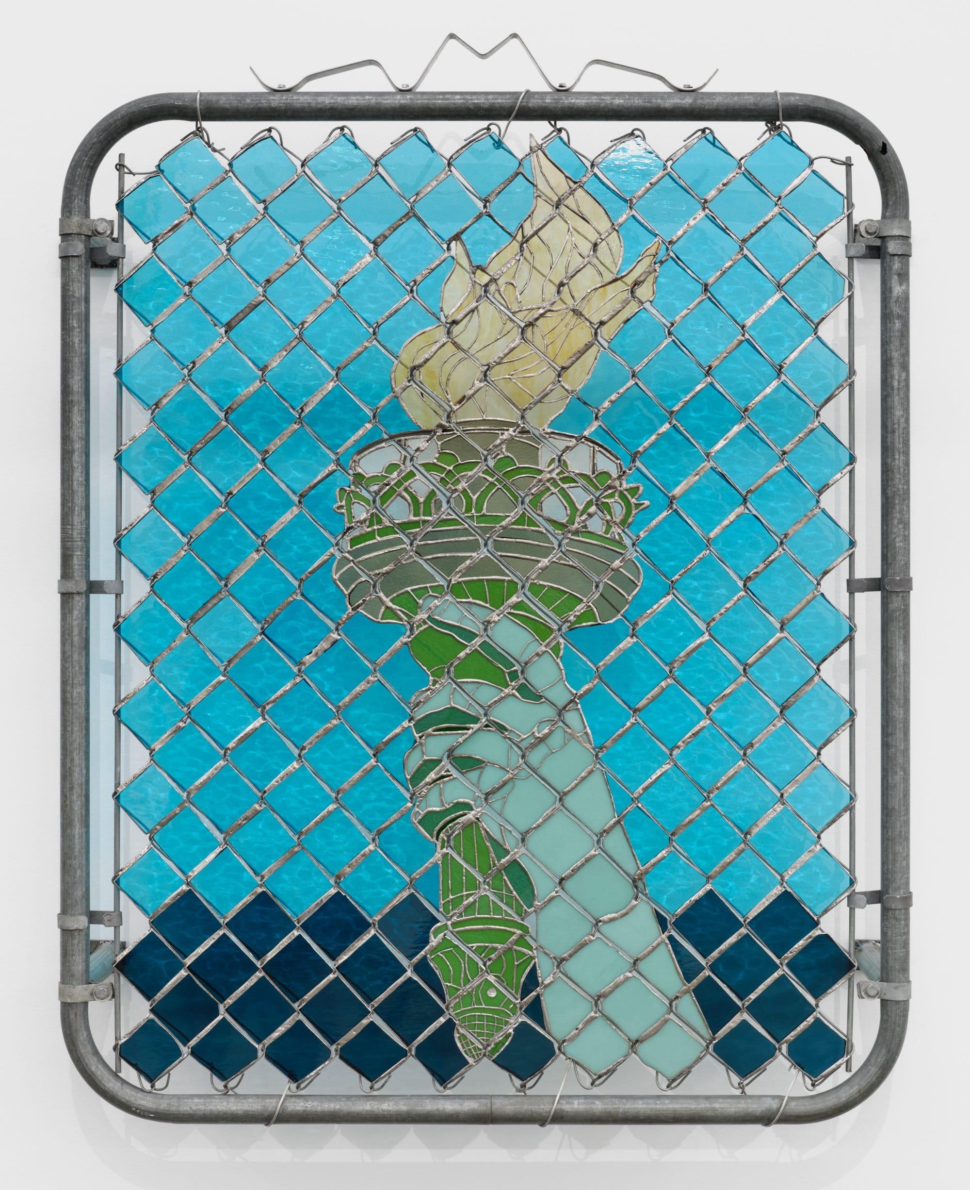 a wall sculpture of stained glass depicting the arm of the Statue of Liberty, installed behind a chainlink gate