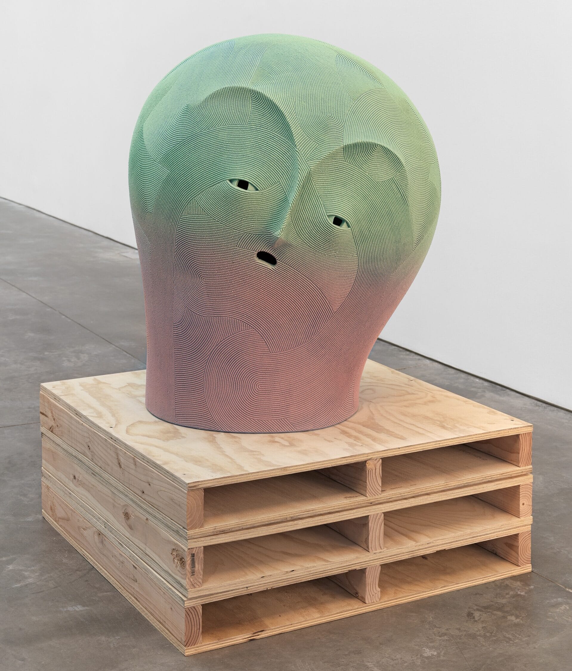 a large ceramic sculpture of a an abstract face in a green-to-pink gradient, installed on a pallet-inspired wood pedestal