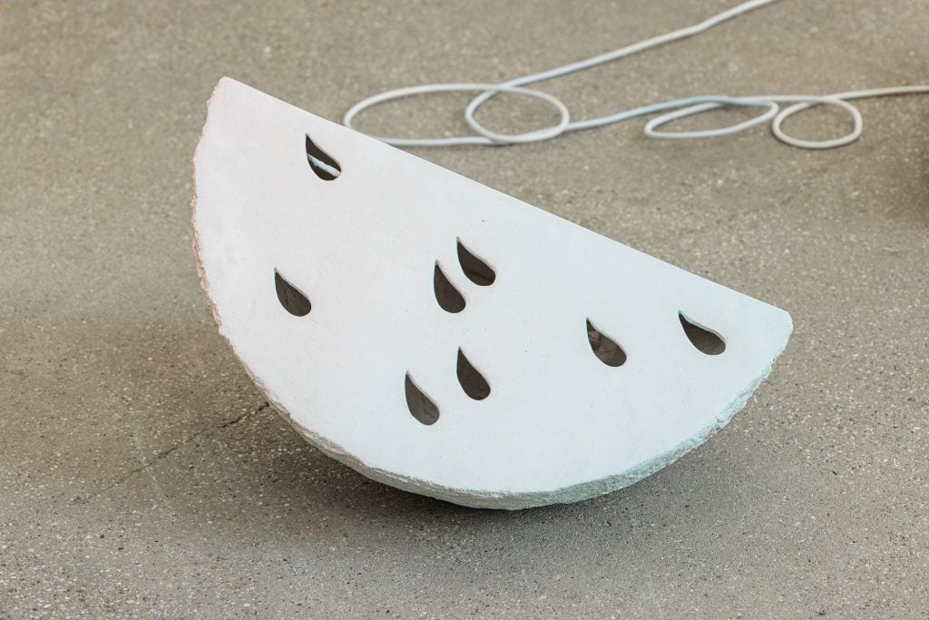 A sculptural artwork titled Fourth Drops by Lukas Thaler, exhibited at Galerie Elisabeth. The piece resembles a minimalist, abstracted slice of watermelon, crafted from a white, stone-like material with a rough, unfinished edge. The surface features teardrop-shaped cutouts in a symmetrical pattern, evoking watermelon seeds. The sculpture rests on a concrete floor, with an electrical cord looping in the background, suggesting an industrial or studio setting.