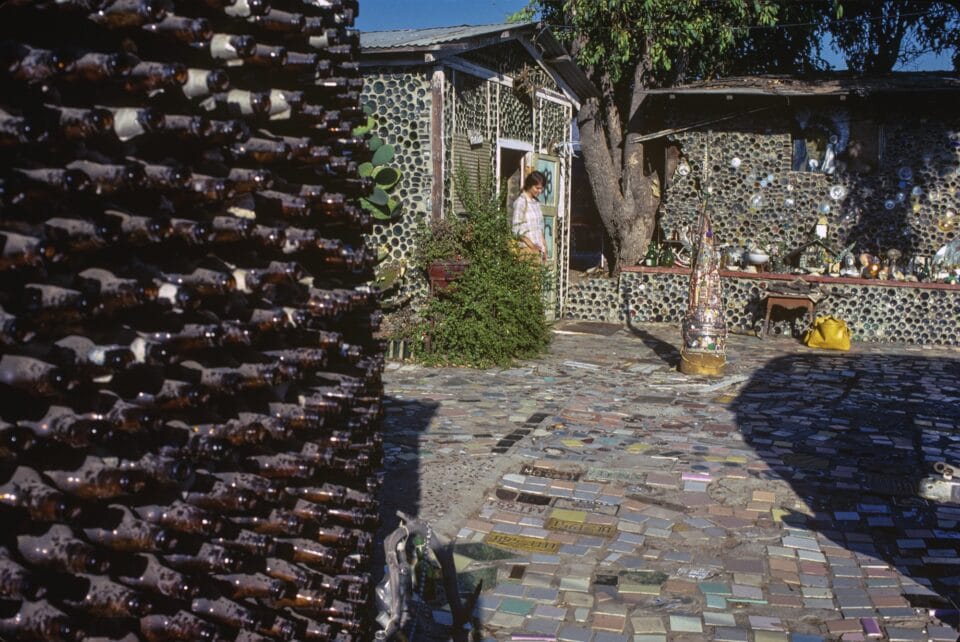 a photograph of a woman known as Grandma Prisbrey outside of her house in California, which is made of glass bottles