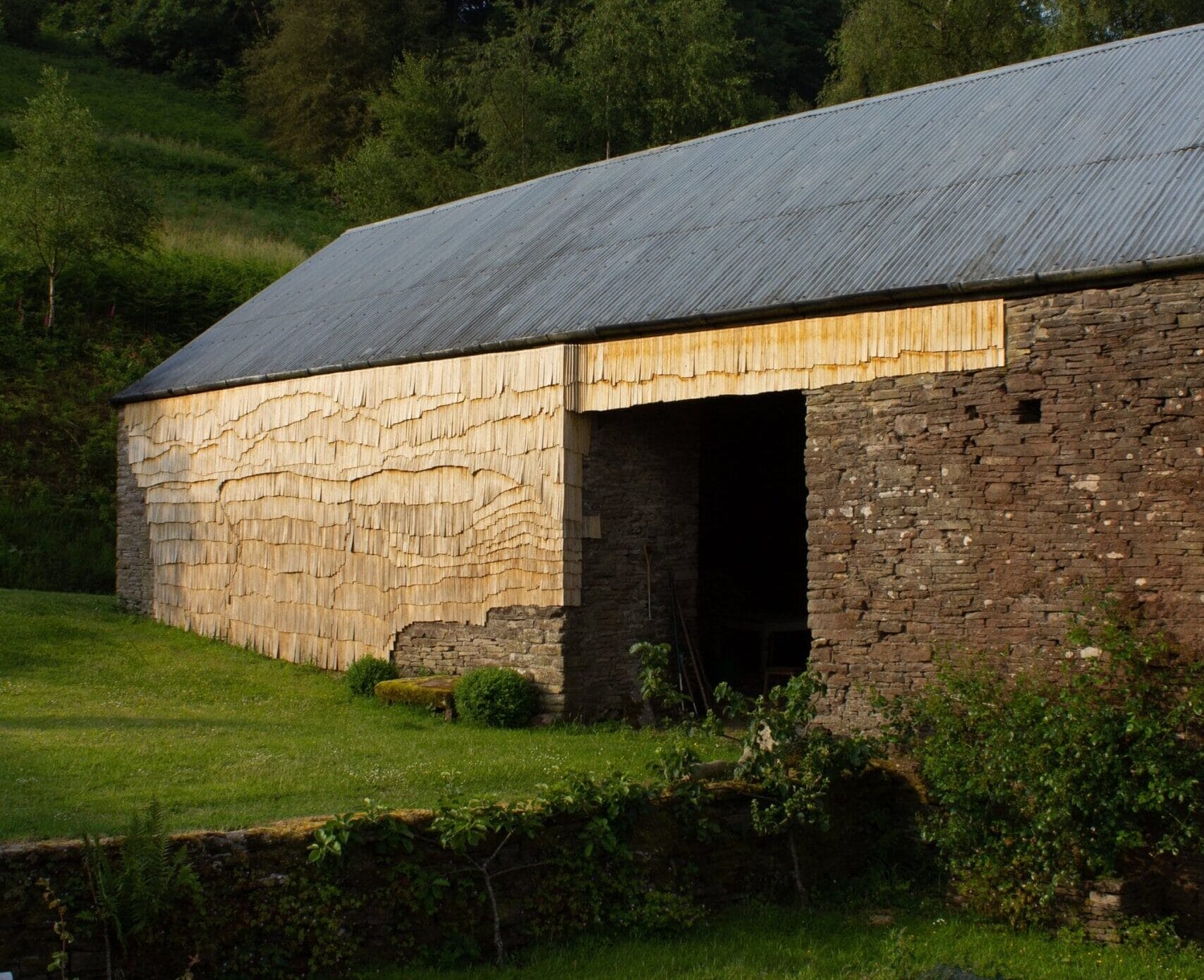 the side of an old stone barn clad in tiny wooden shingles in an undulating pattern