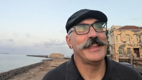 Evan Prodromou Evan Prodromou with a large moustache, smiles in a picture next to the sea.