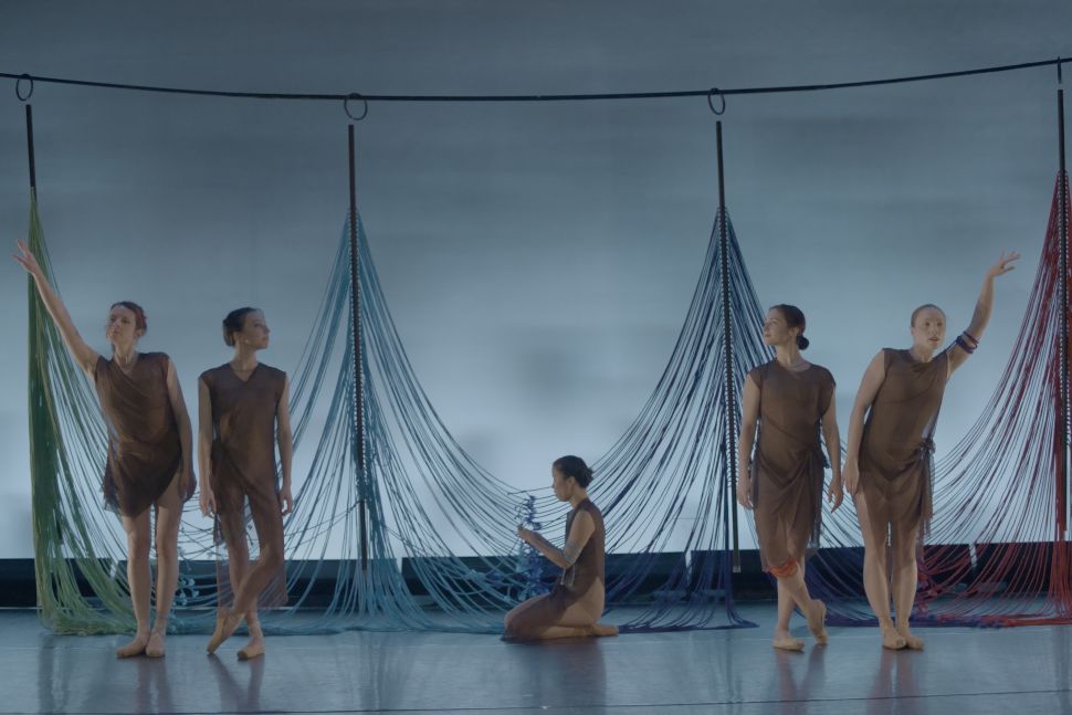 Five ballet dancers dressed in sheer brown costumes performing on a stage, positioned in front of hanging strands of fabric in shades of blue, green and red, evoking a woven or suspended installation.