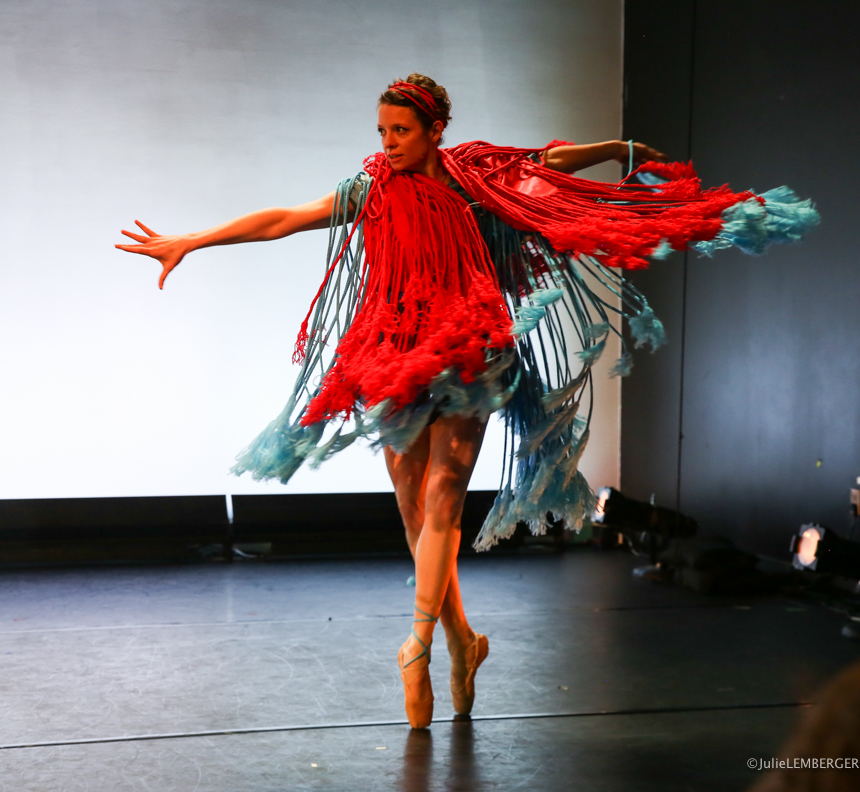 A ballet dancer mid-performance wearing a striking red and blue fringed costume, extending one arm dramatically while balancing on pointe against a minimal, light-colored stage background.