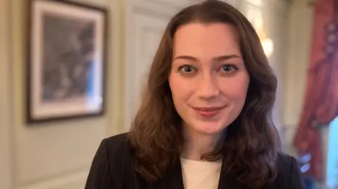 Mousumi Bakshi/BBC Dr Ally Louks has shoulder length brown hair, blue eyes, and is smiling at the camera. She is wearing a black blazer and white t-shirt. She is standing in a room and the lighting is casting a warm glow.