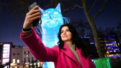 PA Media A female member of the public takes a selfie in front of a large, inflatable blow-up blue cat