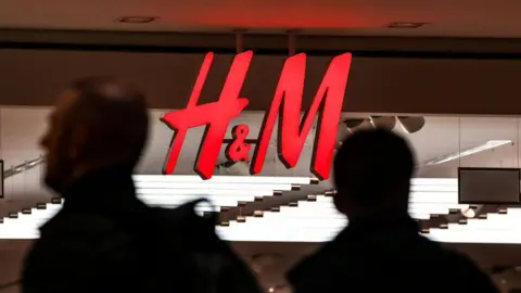 Getty Images Bright red H&M logo displayed on a store sign, with the silhouettes of two passers-by walking past underneath it. 