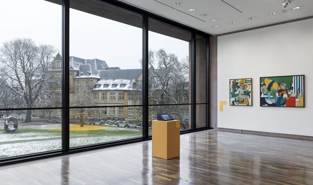 an installation view of two paintings on a gallery wall with large windows overlooking a wintry courtyard to the left