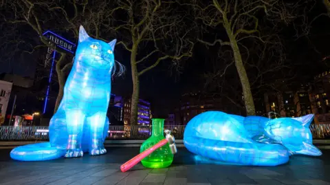 Art of London/PA Media Towering five-metre (16ft) neon inflatable cats, coloured bright blue, are in Leicester Square, the size of a double-decker bus. They are pictured at night.