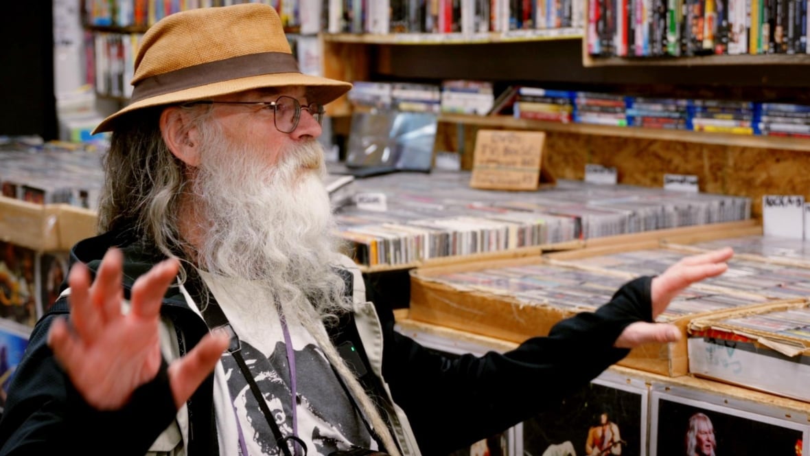 Man with a white beard and straw hat sits in front of a stack of cassette tapes. 