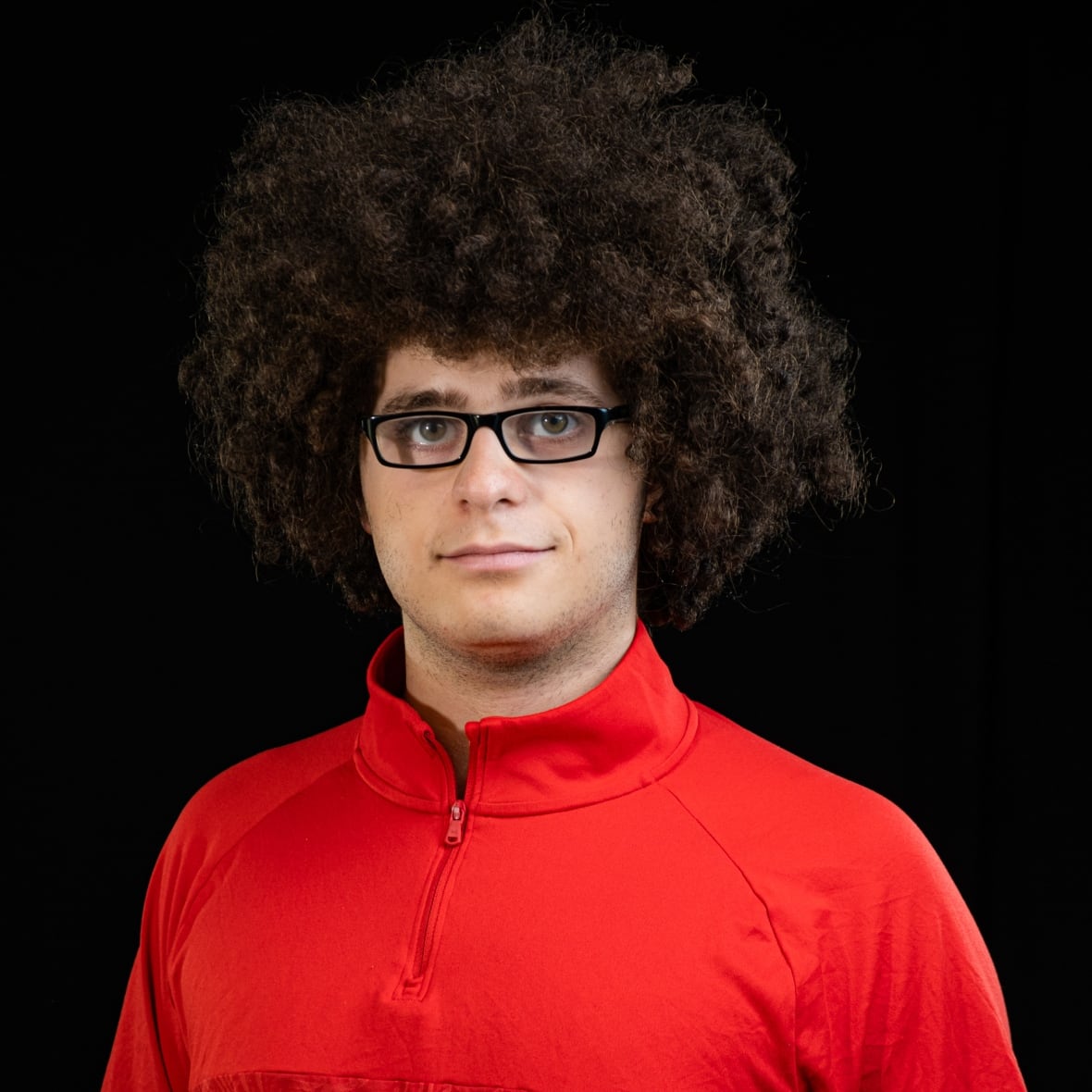 Young man with big, brown curly hair and glasses has a slight smile. He is wearing a red fleece top. 