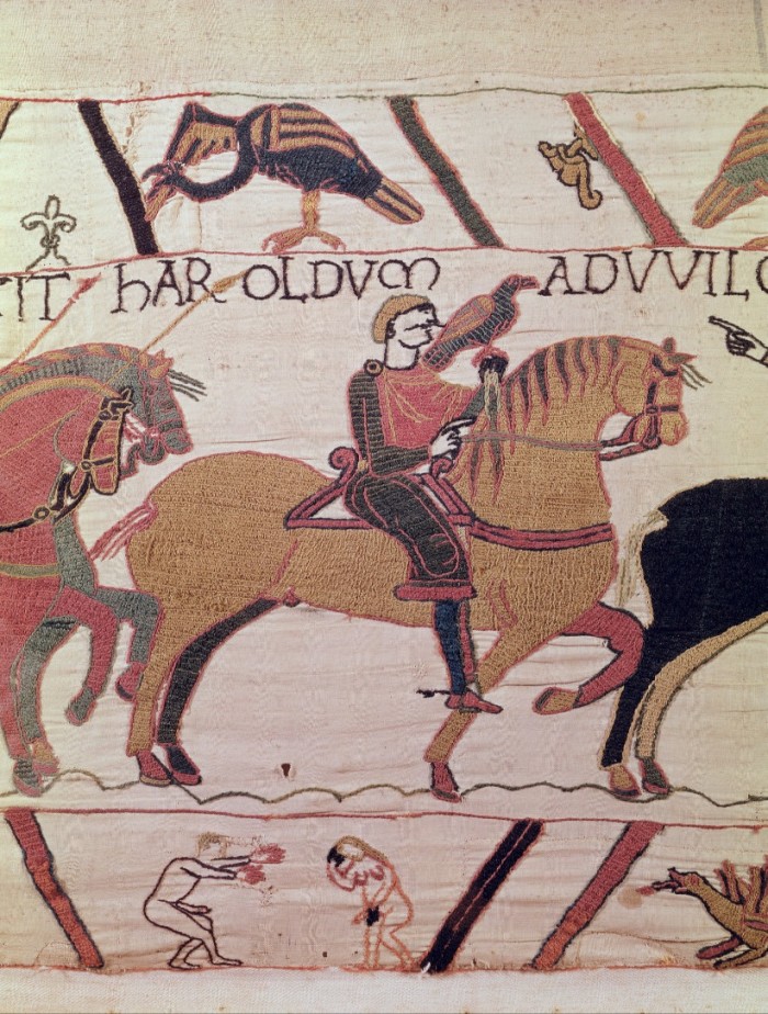 A tapestry of a man on a horse with a bird on his hand. There are other smaller images including a bird, words and a naked man with his hands outstretched towards a naked woman