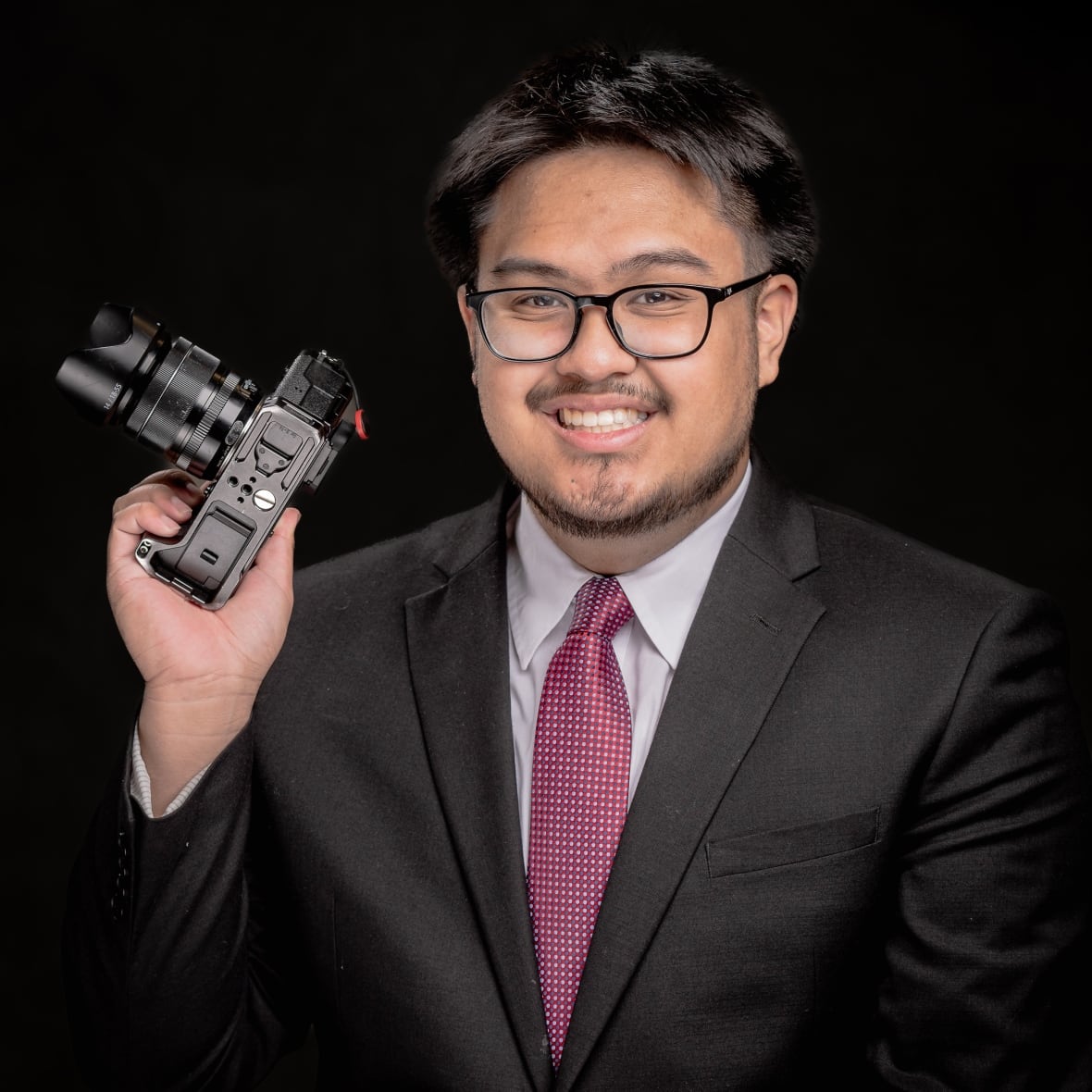 Young man with thin, black-framed glasses holds a camera and smiles in this head and shoulder portrait. He is wearing a dark grey suit, with a white dress shirt and a  light red tie with white checks on it. 