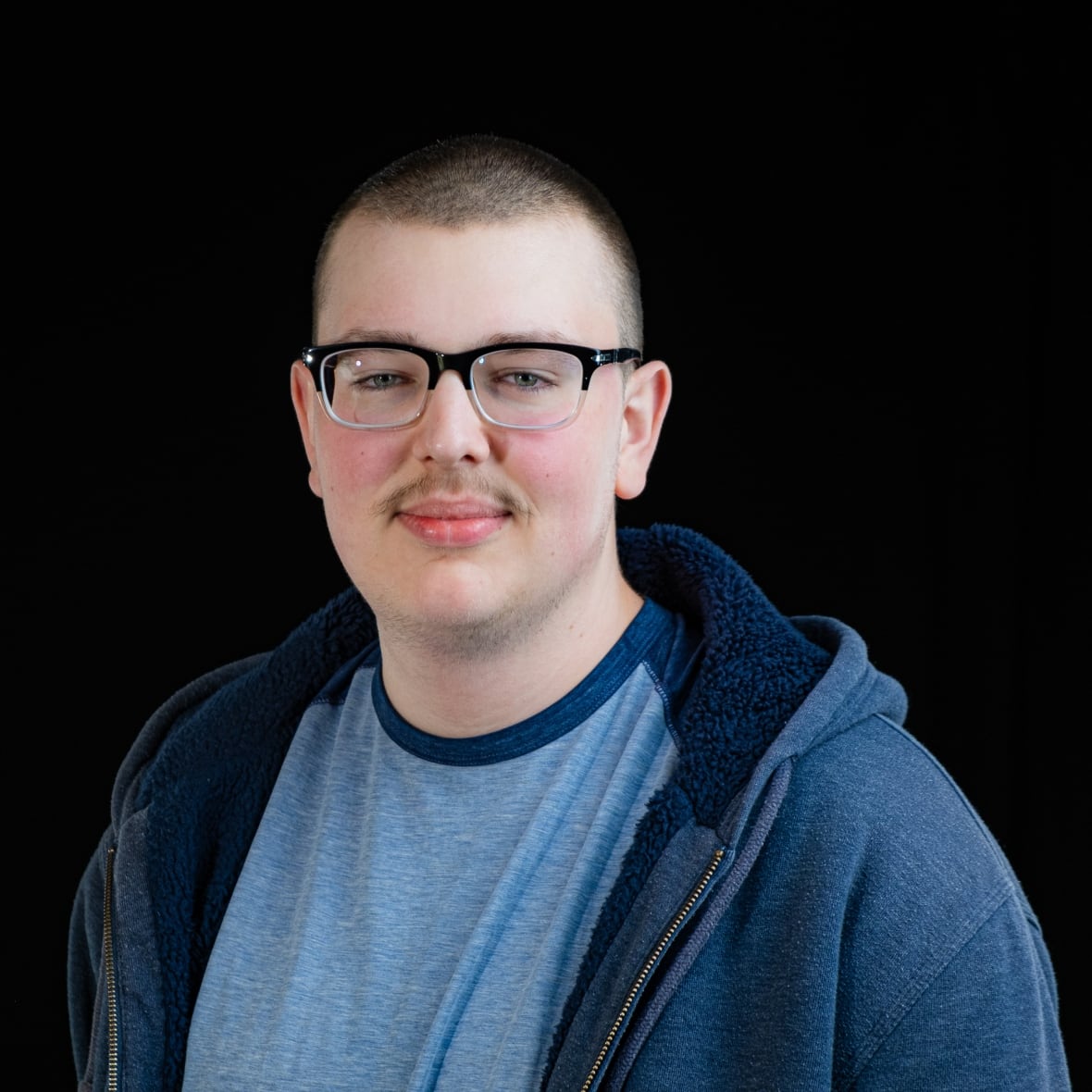 Young, Caucasian-appearing man with black-frame glasses and a small moustache smiles without teeth in this portrait. He is wearing a light blue T-shirt with an unzipped navy blue hoodie over top. 