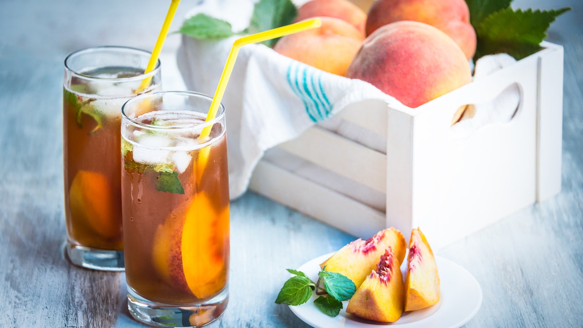 Two glasses of peach-flavored iced tea with straws sit next to some peaches.
