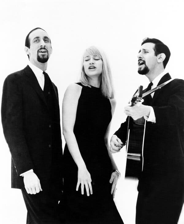 A black-and-white photo of a singing trio, with one group member playing a guitar.