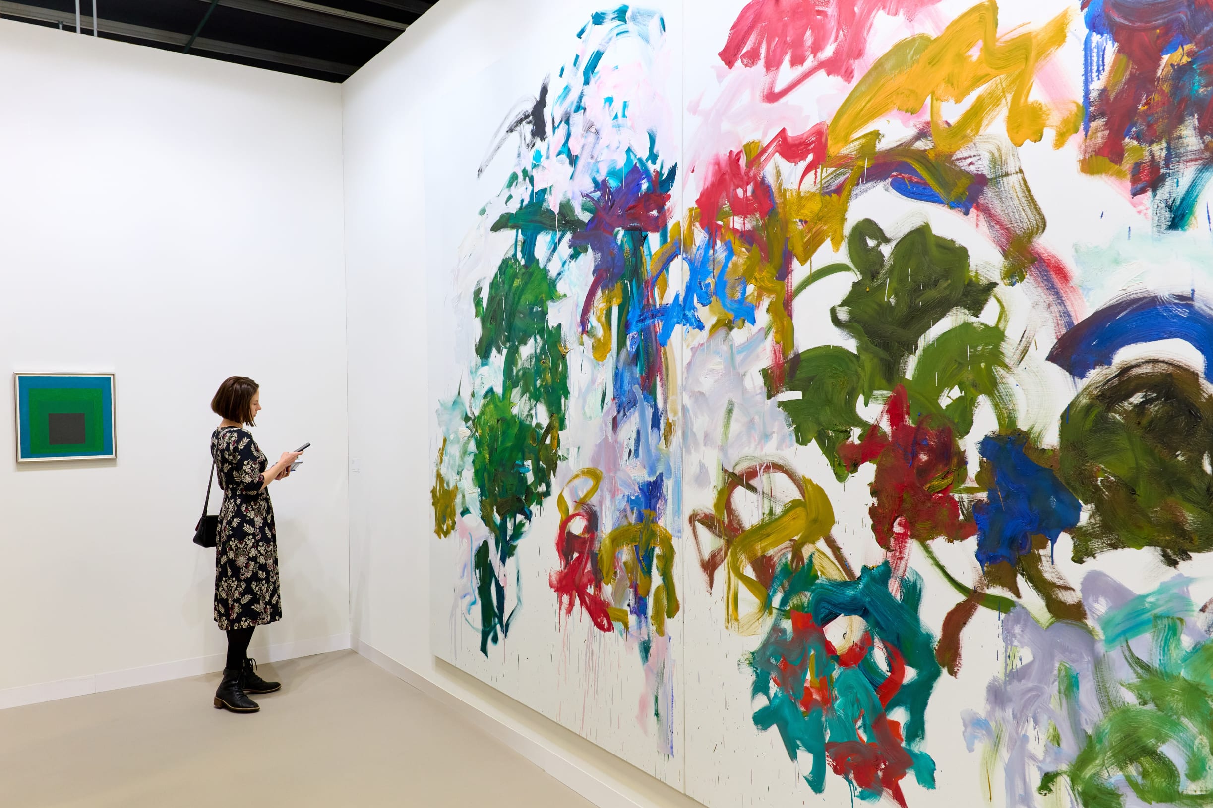 A visitor stands in a gallery looking at a large, colorful abstract painting by Joan Mitchell on David Zwirner's booth wall, with a smaller geometric painting on an adjacent wall.