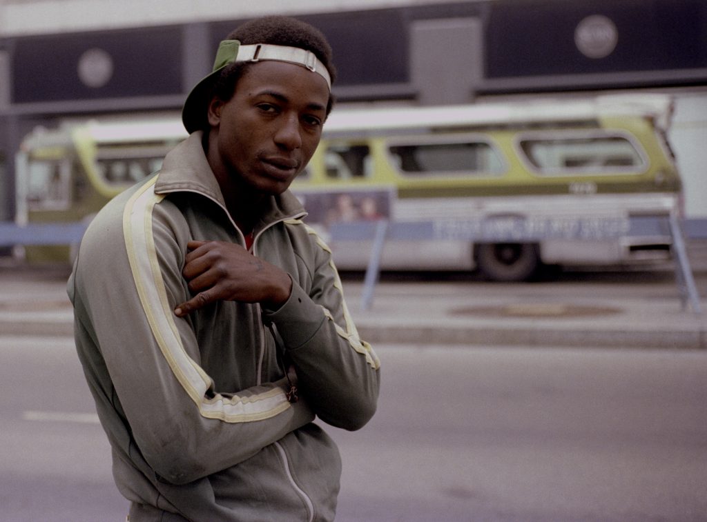 Man wearing track jacket posing on street in front of bus.