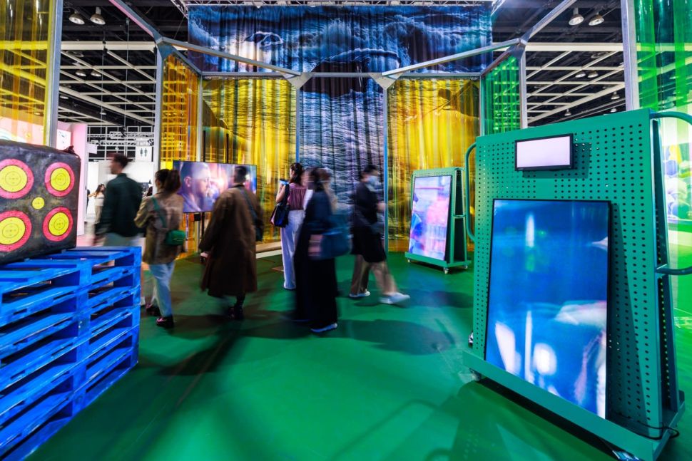 Visitors interact with a colorful, immersive art installation featuring large digital screens, translucent yellow and green plastic curtains, and vivid blue pallets at Art Basel Hong Kong 2025. A video portrait is displayed on one screen, while others show abstract visuals. The space is set on a green floor with overhead industrial lighting, and people are seen walking through and observing the artworks.