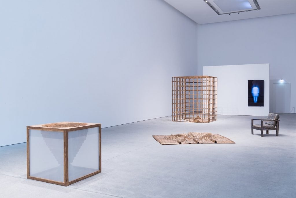 A contemporary art installation in a minimalist gallery, featuring wooden structures, a chair, and a digital screen displaying a glowing figure.